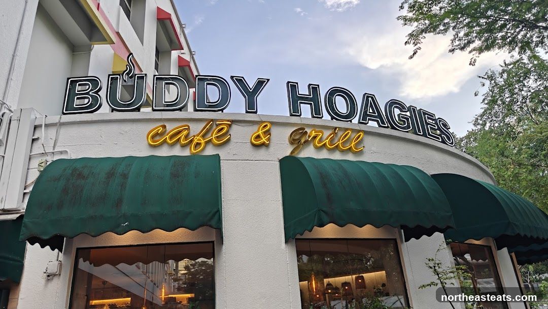 A close-up exterior shot of the "Buddy Hoagies cafe & grill" storefront. The sign is illuminated with neon lights. Green awnings cover the windows below.
