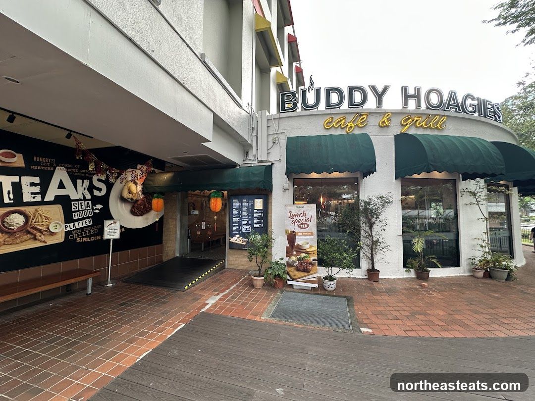 The exterior of a cafe and grill with a large mural on the left advertising steaks, seafood, and platters. The cafe's name, "BUDDY HOAGIES cafe & grill," is prominently displayed above the entrance. Green awnings cover the windows. A lunch special poster is visible on the right.