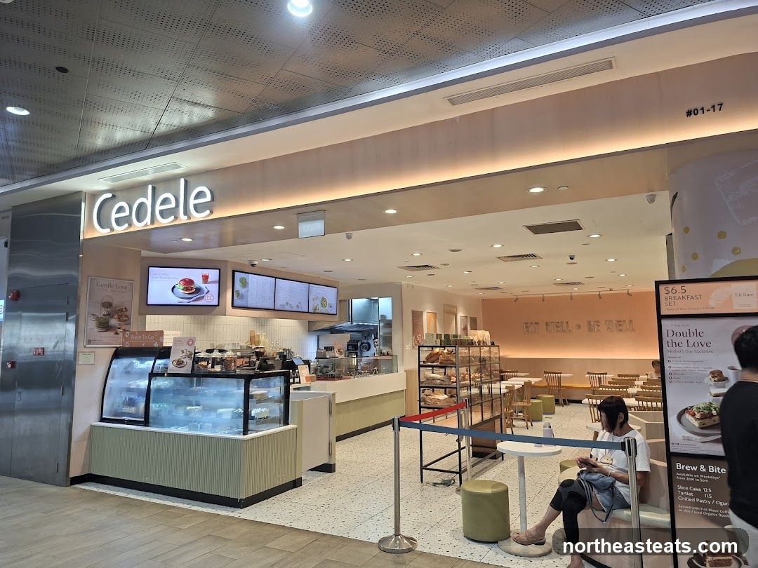 A bright and airy cafe with a display of pastries and cakes. The cafe has a modern design with light wood accents and pastel colors. The brand name "Cedele" is prominently displayed above the entrance.