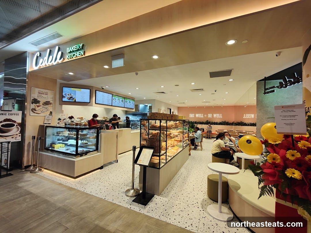 The exterior of a Cedele Bakery Kitchen with a display of cakes and pastries. The shop has a modern design with light wood accents and a speckled floor. There are tables and chairs visible inside, and customers are seated. A sign on the wall reads "EAT WELL • BE WELL".