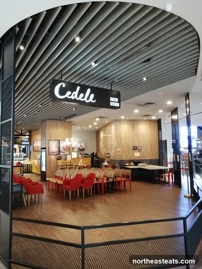 The exterior of a Cedele Bakery Kitchen with a prominent sign. The shop has a modern interior with wooden walls and a slatted ceiling. There are several red chairs and tables arranged in the seating area. A counter with food displays is visible in the background.
