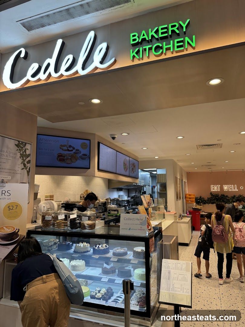 The exterior of a Cedele Bakery Kitchen with a prominent sign. Inside, cakes are displayed in a refrigerated case, and customers are browsing. A staff member is visible behind the counter.