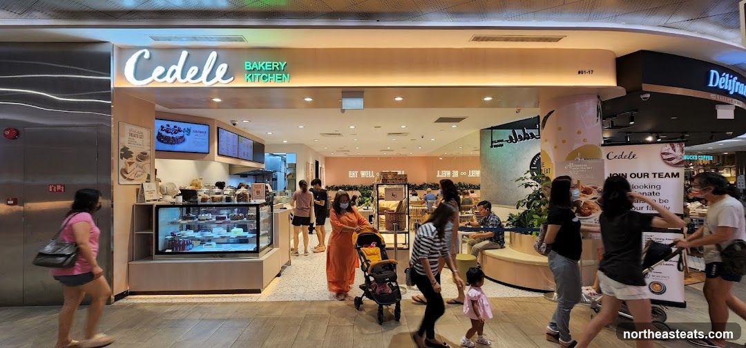 The exterior of a Cedele Bakery Kitchen with people walking by. The storefront features the Cedele logo and "Bakery Kitchen" text. Inside, cakes and pastries are displayed in a refrigerated case. A sign that says "Join Our Team" is visible.