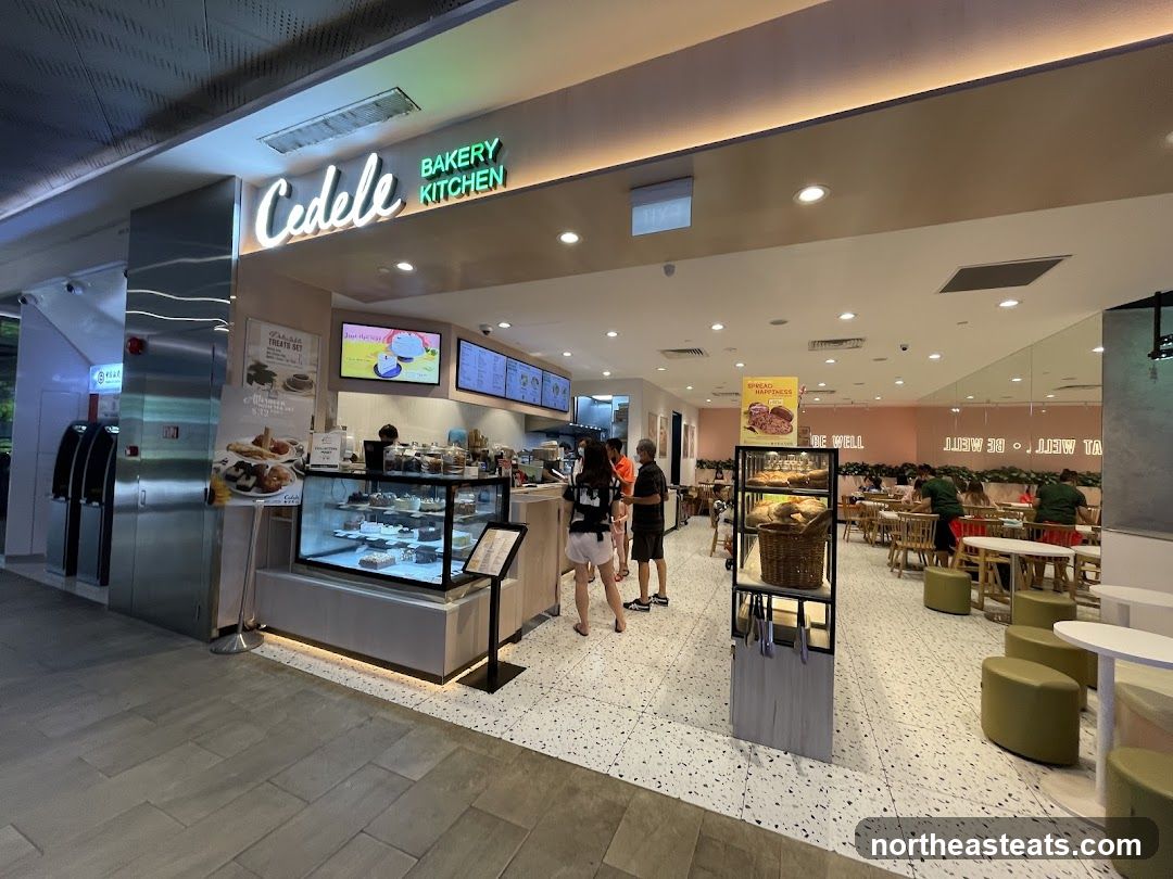 The exterior of a Cedele Bakery Kitchen with a display of cakes and pastries. The shop has a modern design with bright lighting and seating areas visible inside.