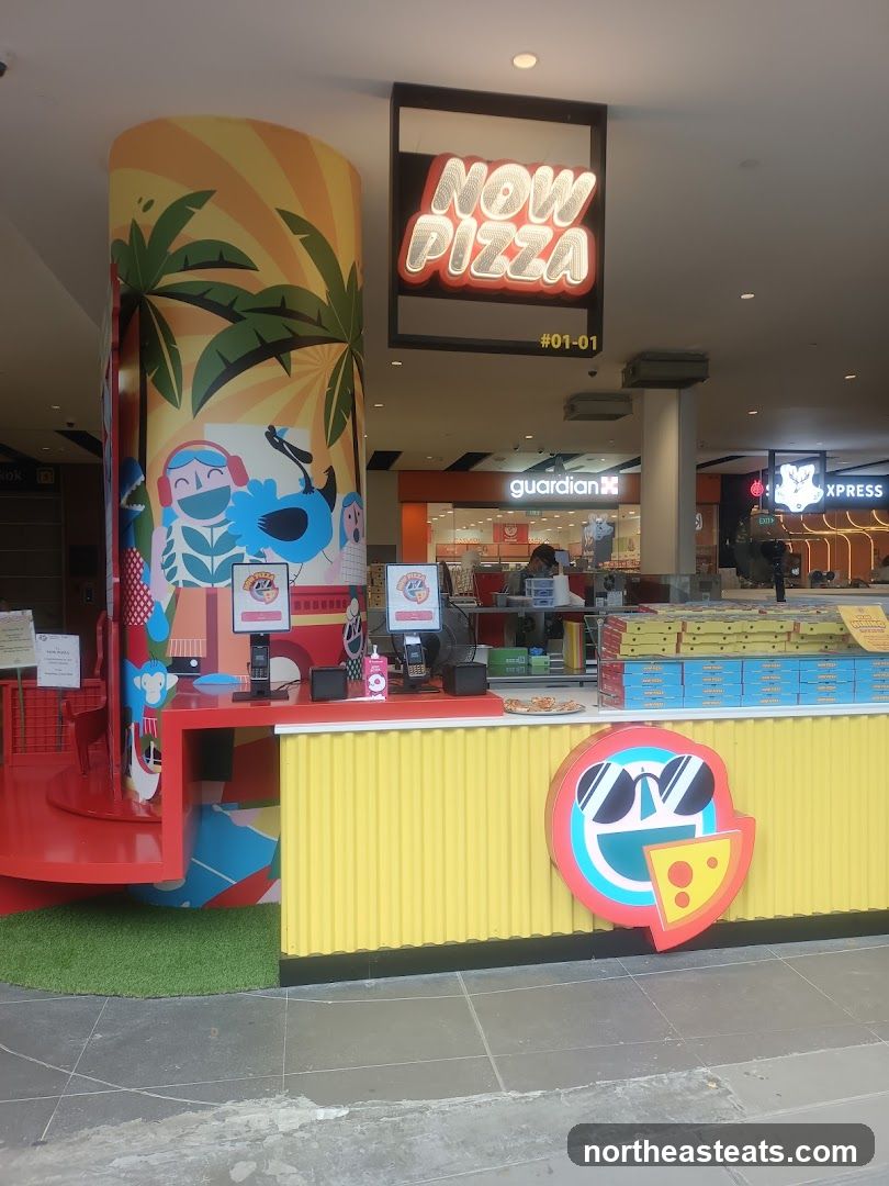 A brightly colored pizza stall with a large sign that says "NOW PIZZA". The stall has a yellow corrugated facade and a red counter. There are two screens on the counter, likely for ordering. A circular logo featuring a slice of pizza with sunglasses is visible on the front. Behind the counter, stacks of pizza boxes are neatly arranged. The stall is located in a mall, with other shops visible in the background.