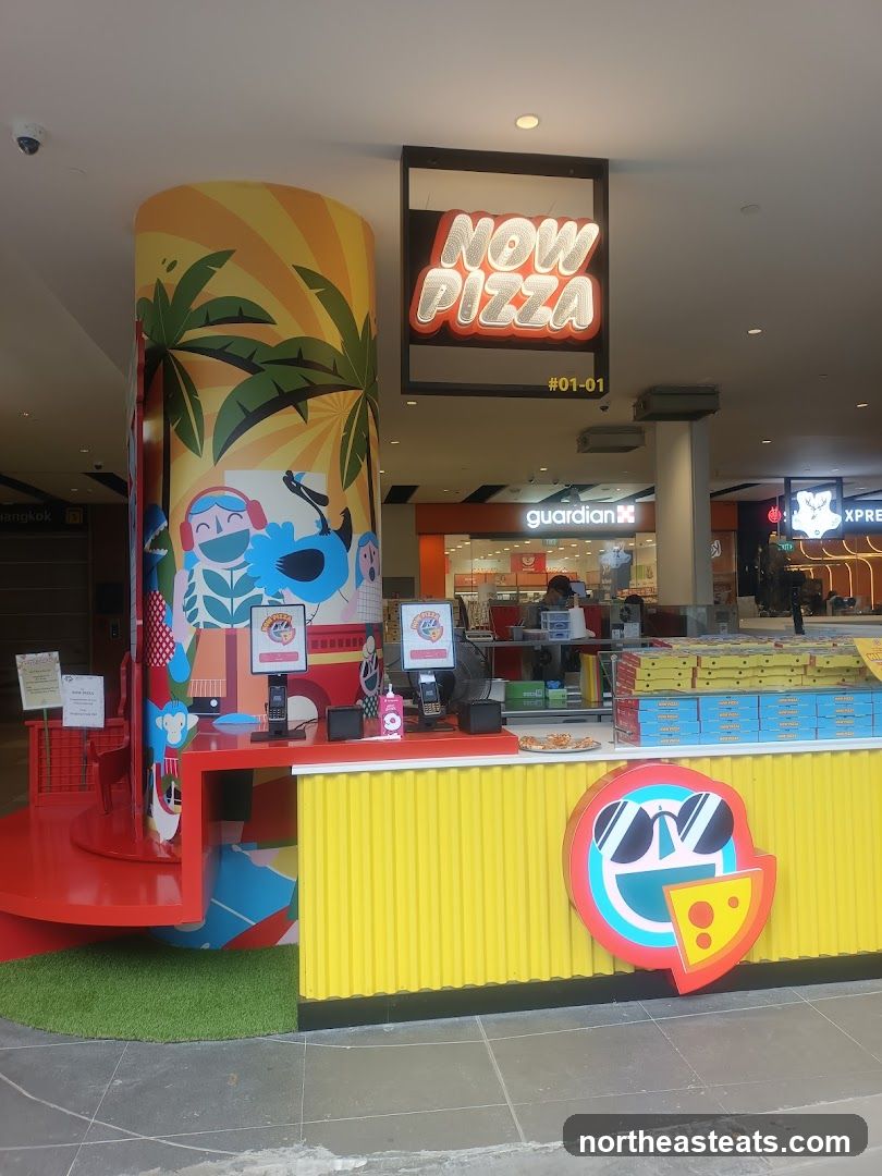 A brightly colored pizza shop with a large, illuminated sign that reads "NOW PIZZA". The storefront features a yellow corrugated facade and a large circular logo with sunglasses and a slice of cheese. A decorative pillar with tropical graphics stands to the left. Pizza boxes are stacked behind the counter.