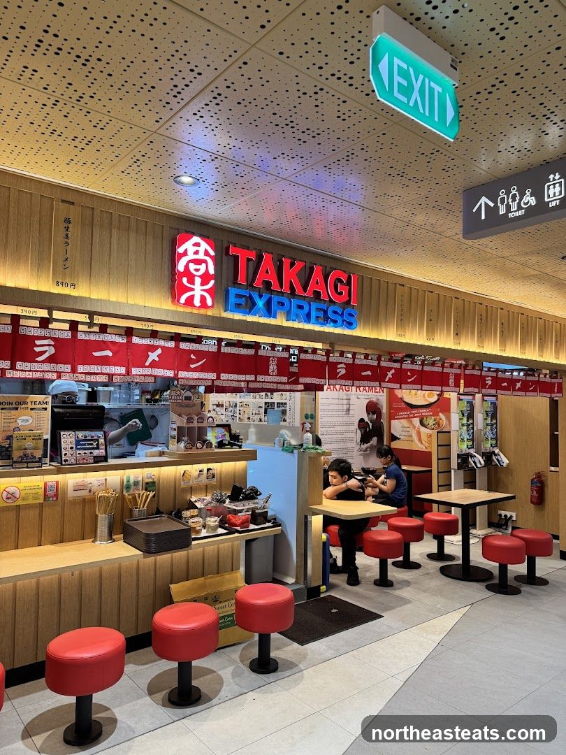 The exterior of a ramen shop with a prominent "TAKAGI EXPRESS" sign. Red banners with Japanese text hang above the counter. Red stools are arranged around small tables. An "EXIT" sign is visible on the ceiling.
