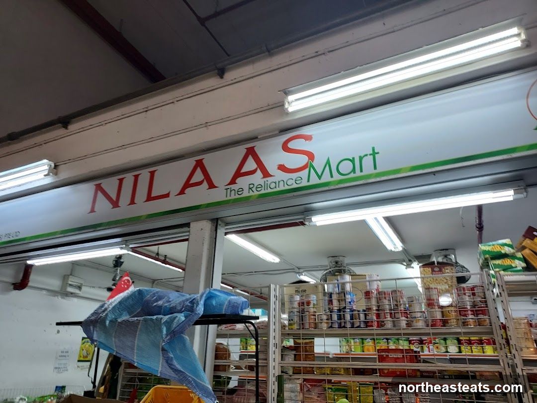 A sign for Nilaas Mart, The Reliance, is visible above a store entrance. The store appears to be a grocery or convenience store, with shelves stocked with canned goods and other items visible inside.