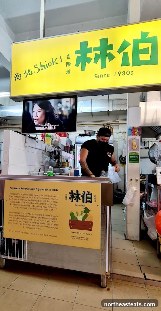 A food stall with a yellow sign featuring Chinese characters and English text that reads "南北Shiok!" and "Since 1980s". Below the sign, a yellow board describes the food as "Authentic Penang Taste Enjoyed Since 1980s" and mentions "Traditional Penang Local Salad" and "Penang Fruit Rojak". A man in a black t-shirt and cap is visible behind the counter, preparing food. A television screen above the counter shows a woman speaking.