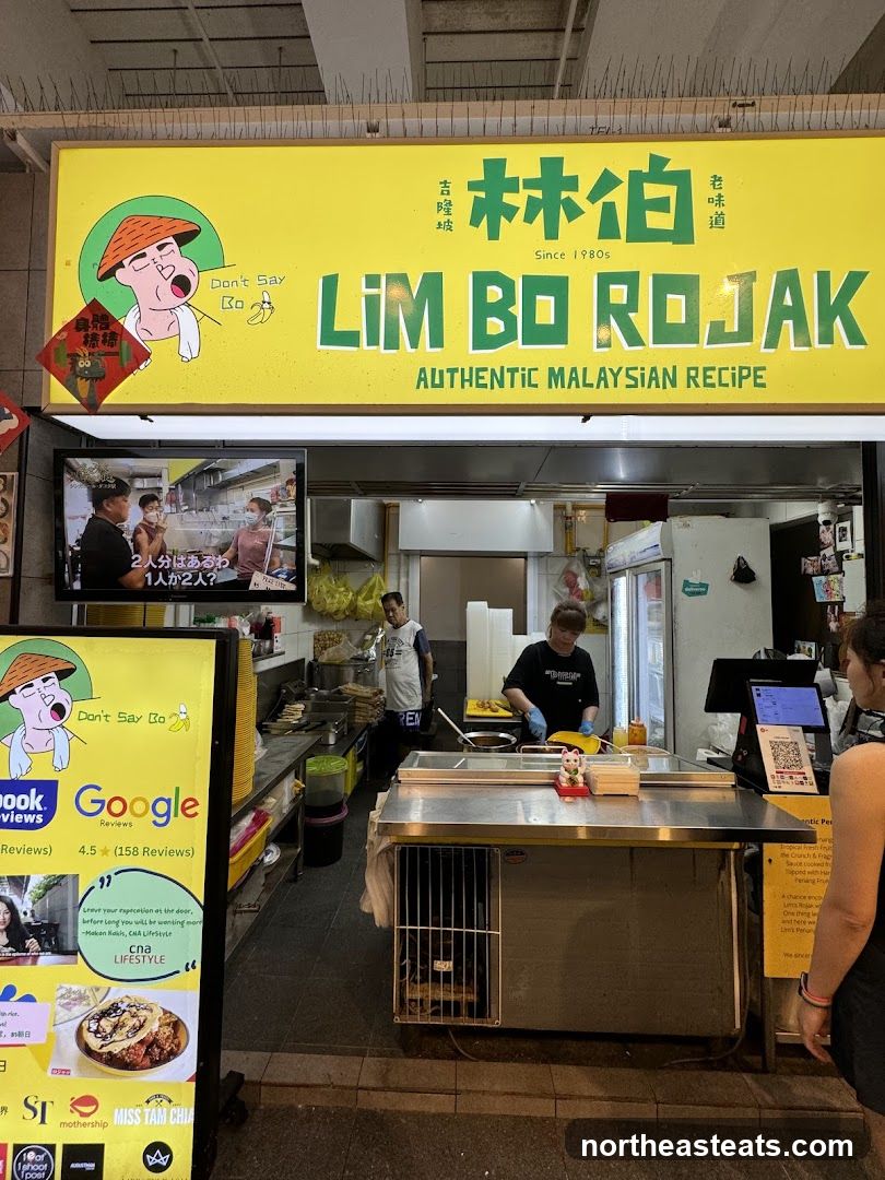 A street food stall with a bright yellow sign that reads "LIM BO ROJAK AUTHENTIC MALAYSIAN RECIPE" in large green letters. Below the sign, the stall is open, showing staff preparing food. A television screen on the left displays a video with Japanese subtitles. A yellow stand with reviews and images of food is also present.