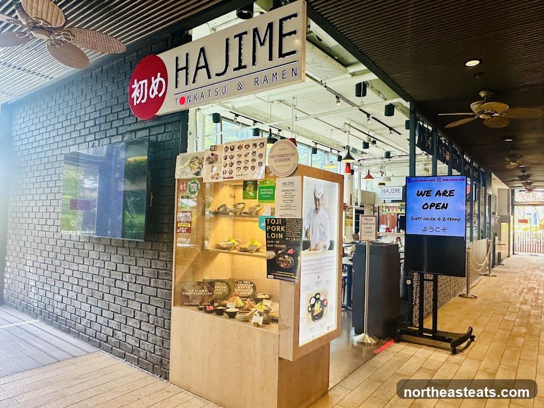 The exterior of a restaurant called Hajime, specializing in Tonkatsu and Ramen. A large sign above the entrance reads "HAJIME TONKATSU & RAMEN". To the left of the entrance is a display case with food samples. To the right, a digital screen displays "WE ARE OPEN" and "ようこそ".
