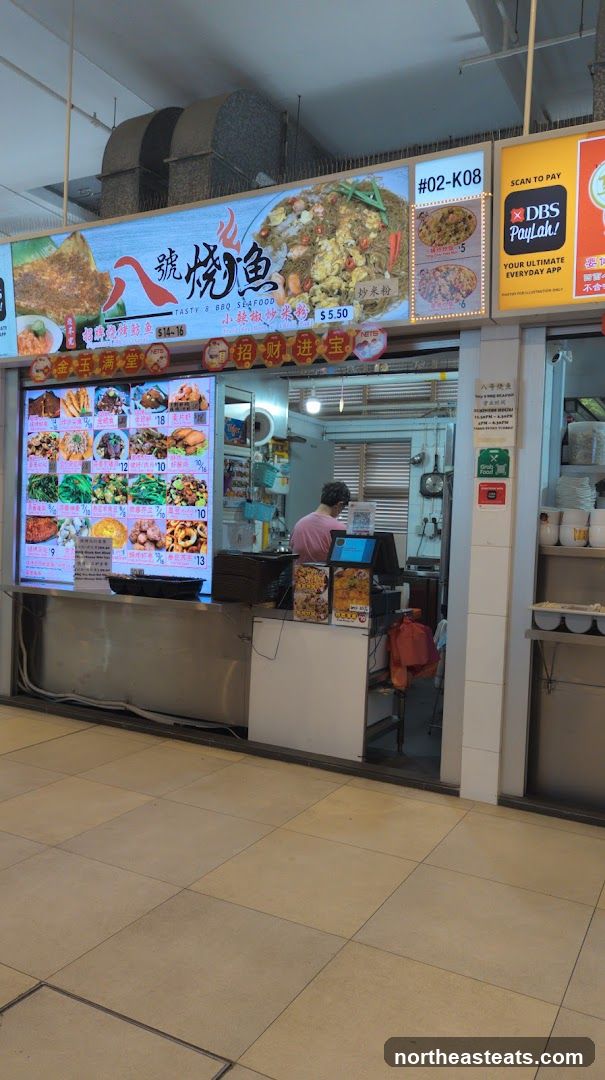 A food stall with a large illuminated menu board displaying various dishes and prices. A person is visible behind the counter. The stall has signage with Chinese characters and English text.