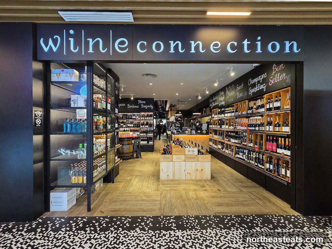 The exterior of a wine shop with the name "Wine Connection" displayed prominently above the entrance. The shop is well-stocked with bottles of wine and spirits on shelves lining the walls. Wooden crates are used for display, and there is a counter area visible in the background.