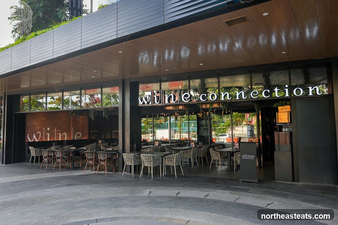 The exterior of a Wine Connection restaurant with outdoor seating. The restaurant has large glass windows and a wooden ceiling. The name "Wine Connection" is displayed in large white letters above the entrance.