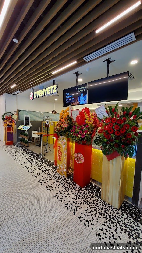 The storefront of D'PENYETZ features a prominent sign with the restaurant's logo. The entrance is decorated with large floral arrangements and balloons, suggesting a celebratory occasion. The floor has a distinctive mosaic pattern of white and black hexagonal tiles. Inside, a counter area with a digital ordering screen and a yellow wall are visible.