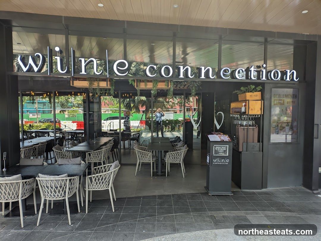 The exterior of a Wine Connection establishment, featuring a prominent sign with the business name. The storefront has large glass windows revealing an indoor seating area with tables and chairs. Outside, there are also tables and chairs set up on the patio. A stand with a menu and a sign indicating 'DEAL OF MONTH' is visible near the entrance.