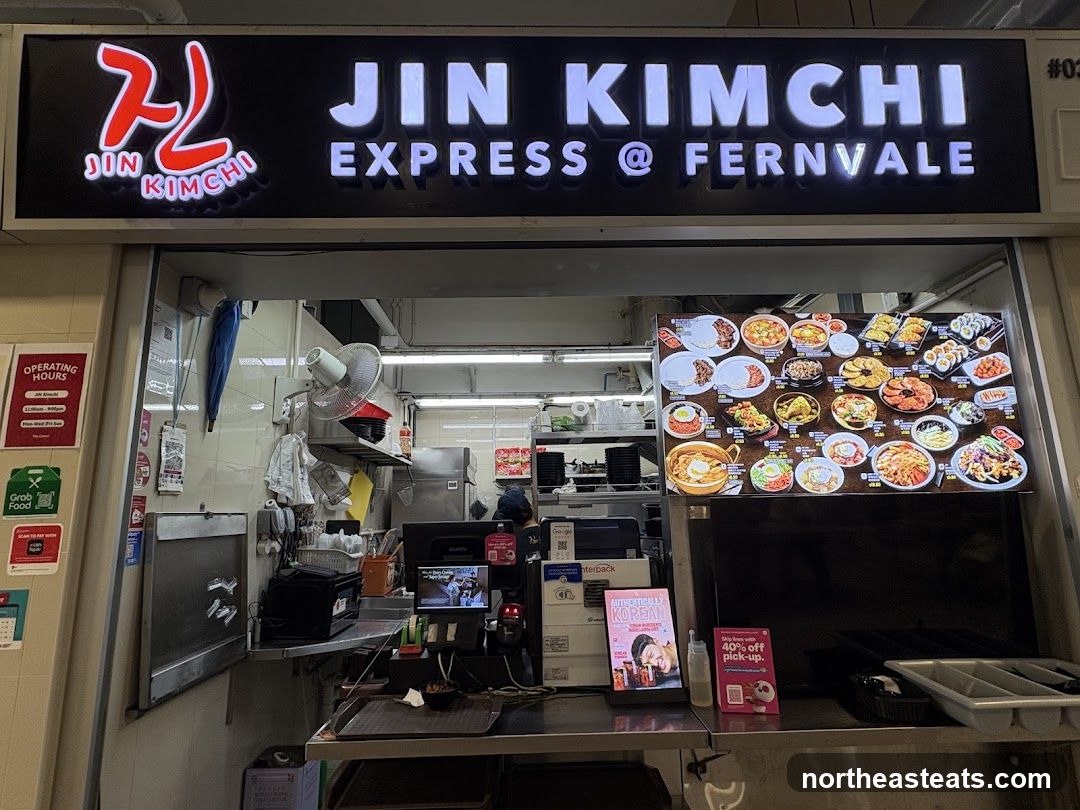 A storefront for Jin Kimchi Express @ Fernvale, a Korean food stall. The stall has a large illuminated sign with the business name and logo. Below the sign is a display of food items and a menu. There are also operating hours posted on the left side of the stall.