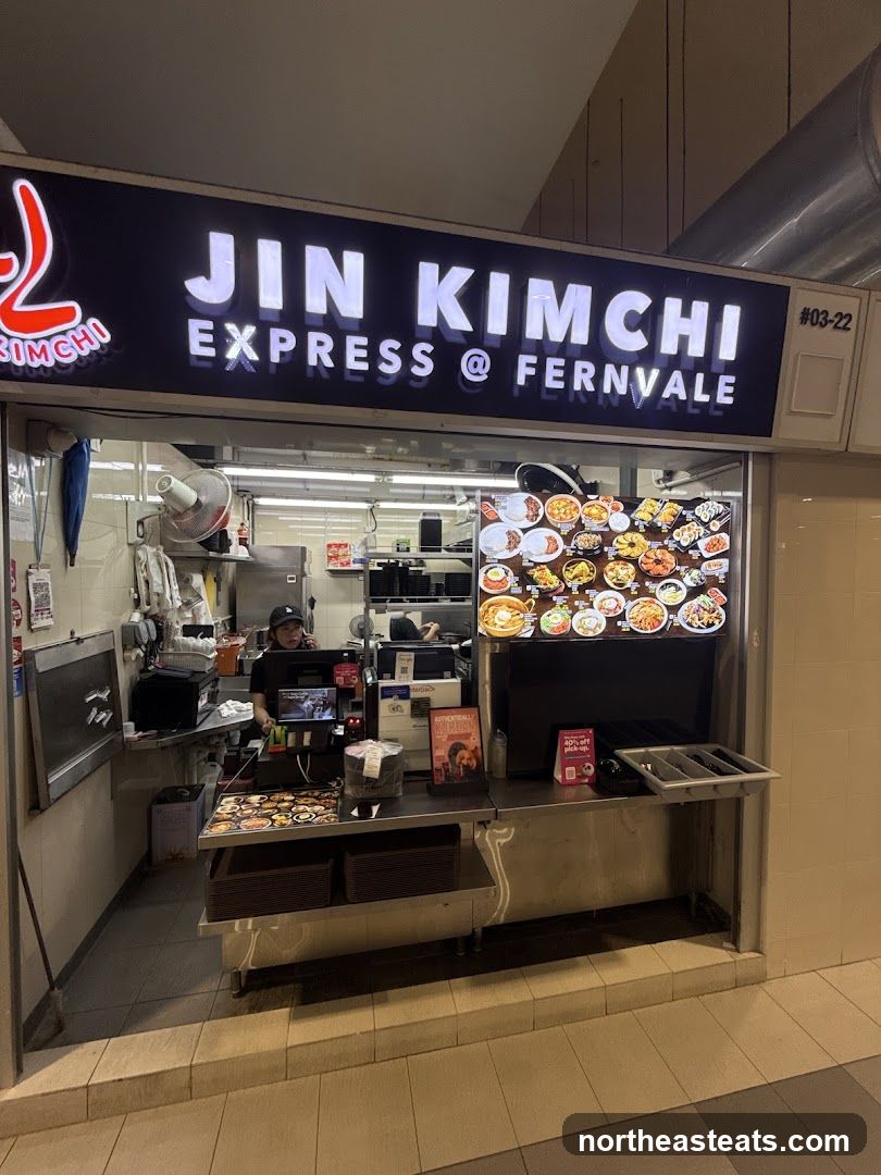 A brightly lit storefront for Jin Kimchi Express @ Fernvale. The signage above the counter displays the business name in large white letters. Below the signage, a digital menu board showcases various Korean dishes with colorful images. A staff member is visible behind the counter, attending to customers. The counter area is equipped with a cash register, order taking station, and storage for food containers. A promotional flyer for "40% off pick-up" is displayed on the counter.