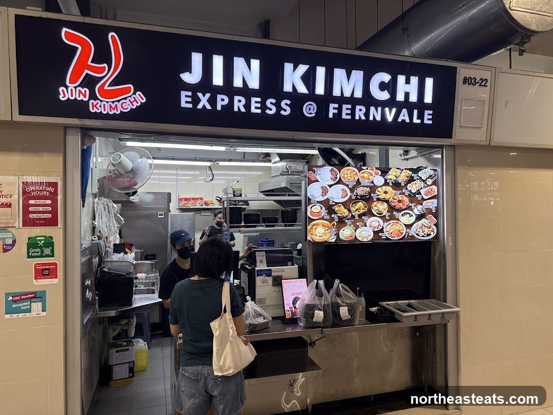 A storefront for Jin Kimchi Express @ Fernvale. The sign above the stall reads "JIN KIMCHI EXPRESS @ FERNVALE" with a logo. Below the sign is a large menu board displaying various Korean dishes. Two staff members are visible behind the counter, and a customer is standing in front of the counter. There are also various signs and posters on the wall to the left of the stall.
