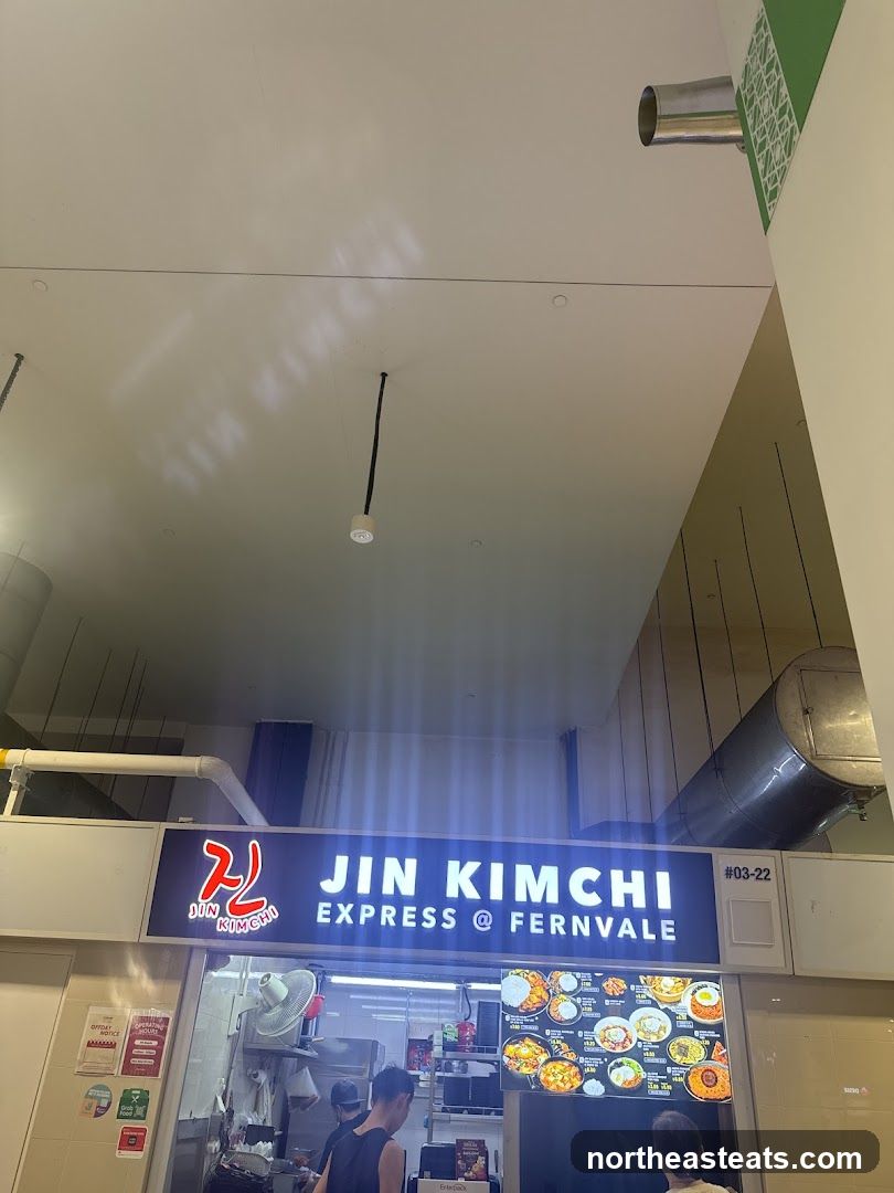 A brightly lit sign for Jin Kimchi Express @ Fernvale is visible above a food stall. Below the sign, a menu board displays various Korean dishes with images and prices. Two customers are visible in front of the stall.