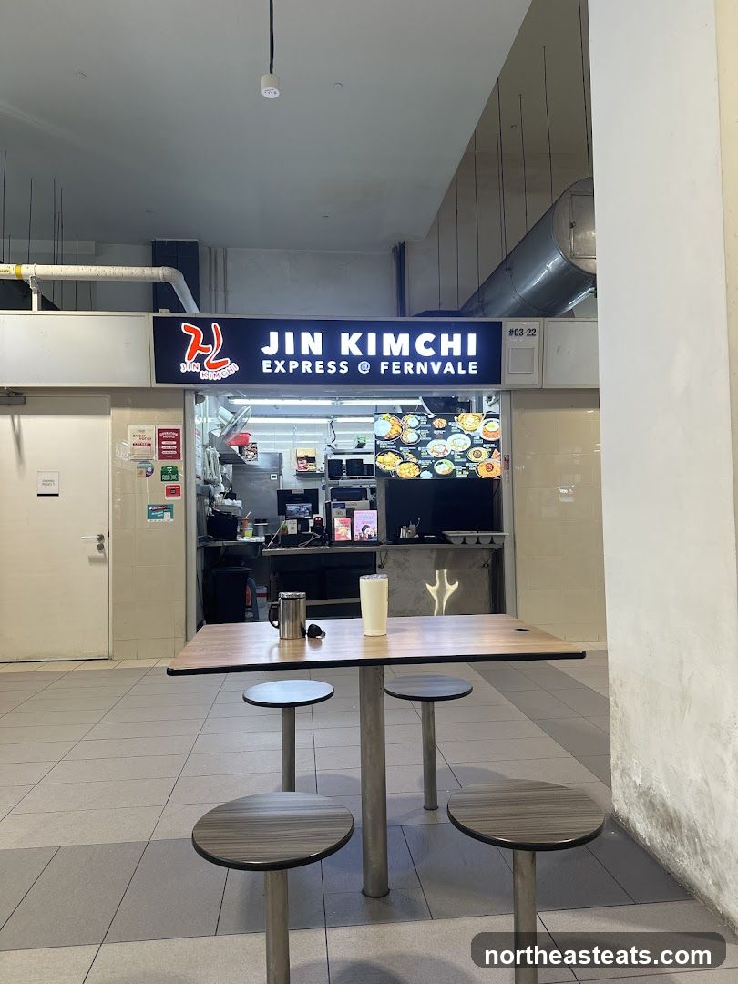 The exterior of a food stall with a prominent sign that reads "JIN KIMCHI EXPRESS @ FERNVALE". The stall appears to be in a food court or hawker center, with a seating area in front. The stall has a display of food items and a counter where orders are taken.
