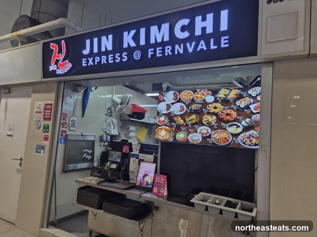 A brightly lit storefront with a large illuminated sign that reads "JIN KIMCHI EXPRESS @ FERNVALE". Below the sign is a display of various Korean dishes, including stews, rice bowls, and sushi rolls. The counter area shows a cash register, stacks of containers, and a small promotional flyer.