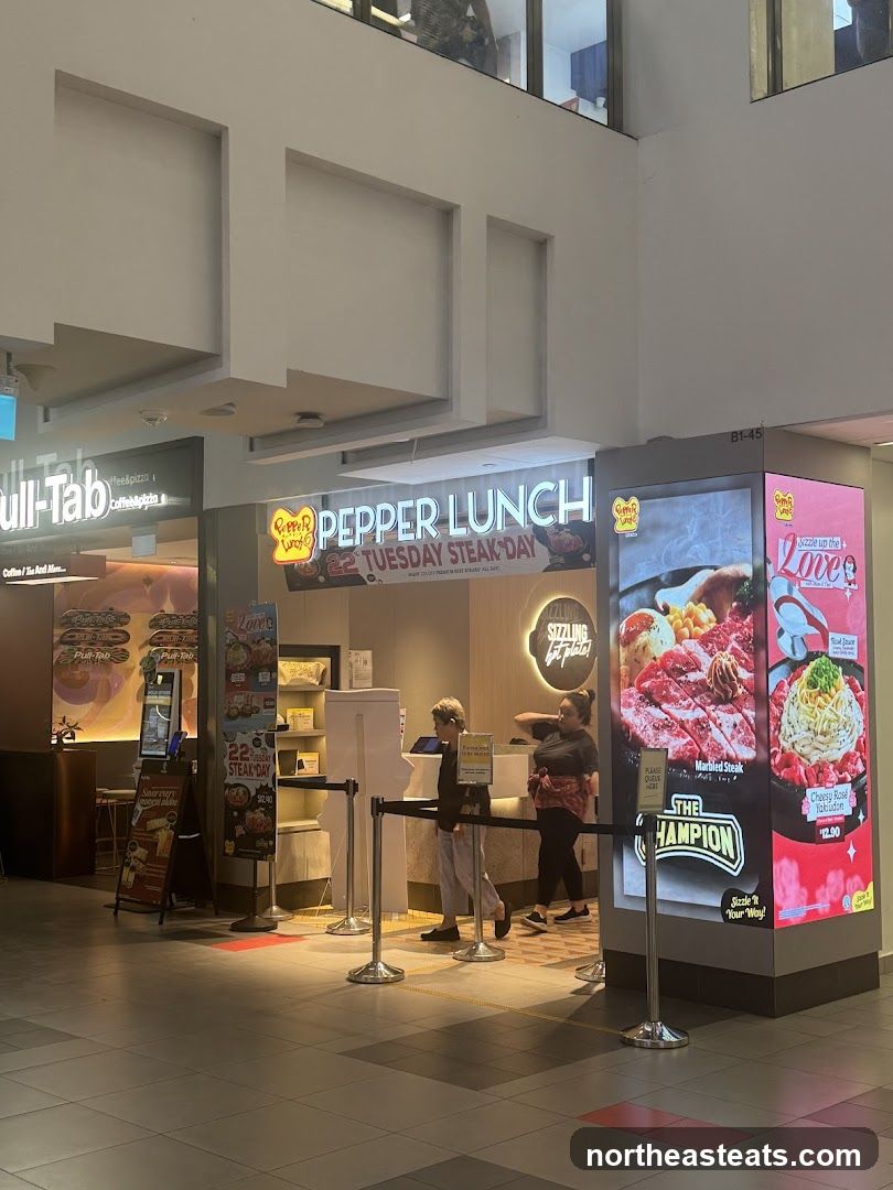 The exterior of a Pepper Lunch restaurant with a prominent sign above the entrance. A promotional display to the right features images of steak and pasta dishes.