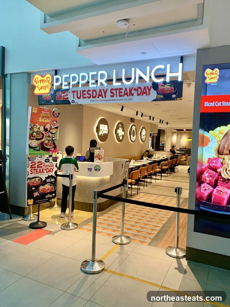The exterior of a Pepper Lunch restaurant with a prominent sign displaying "PEPPER LUNCH" and a promotion for "22nd TUESDAY STEAK DAY". A poster on the left advertises the steak day special with prices. Inside, a counter is visible with staff and customers, and dining tables are set up.