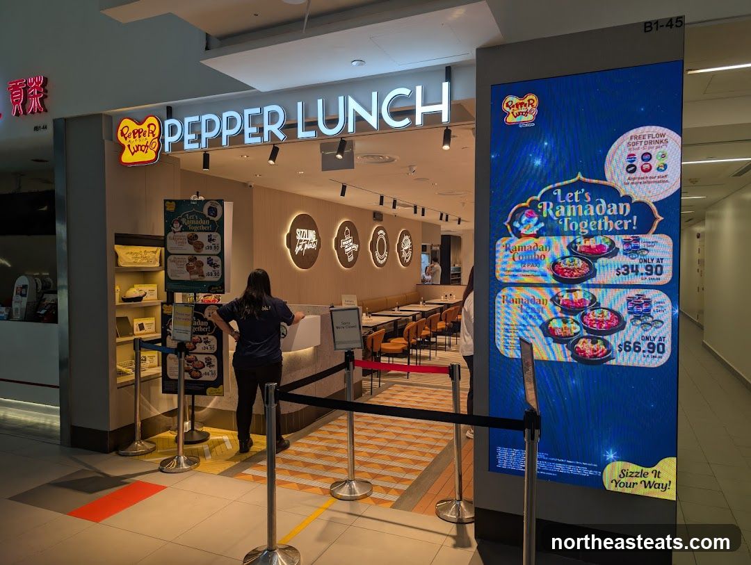 The exterior of a Pepper Lunch restaurant with its prominent signage. A digital display shows a Ramadan promotion. Inside, tables and chairs are visible, along with a counter where a staff member is standing.