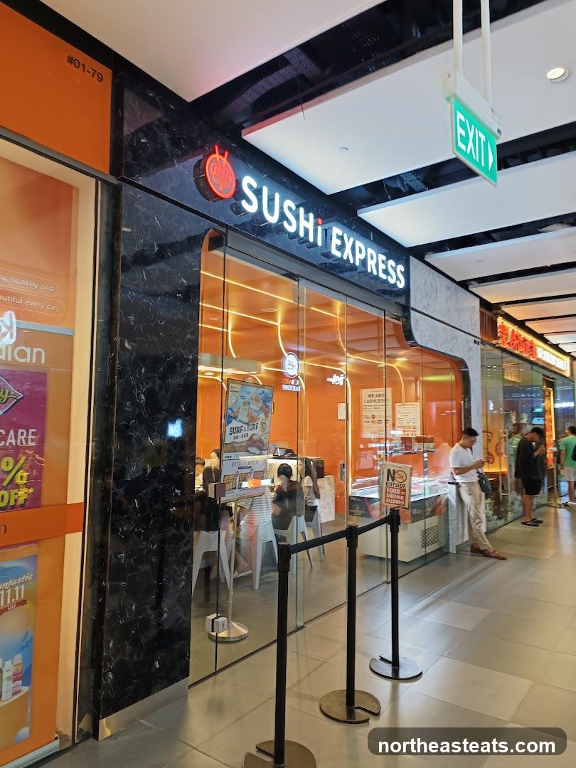 The exterior of a Sushi Express restaurant with a prominent neon sign above the glass storefront. Inside, customers are seated at tables, and there is a display of food items. An "EXIT" sign is visible on the ceiling.