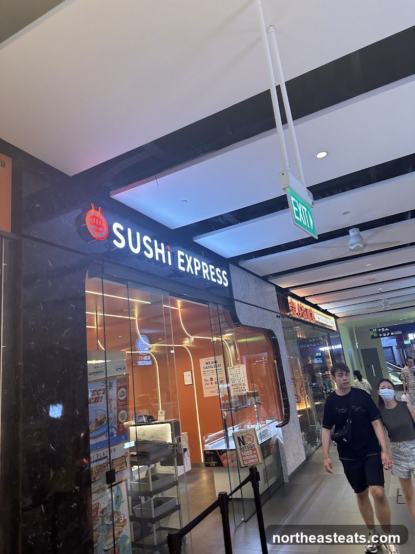 The exterior of a Sushi Express restaurant with a prominent illuminated sign above the entrance. The storefront features large glass windows, revealing the interior with orange walls and display cases. A sign indicating "WE ARE CASHLESS" is visible inside.
