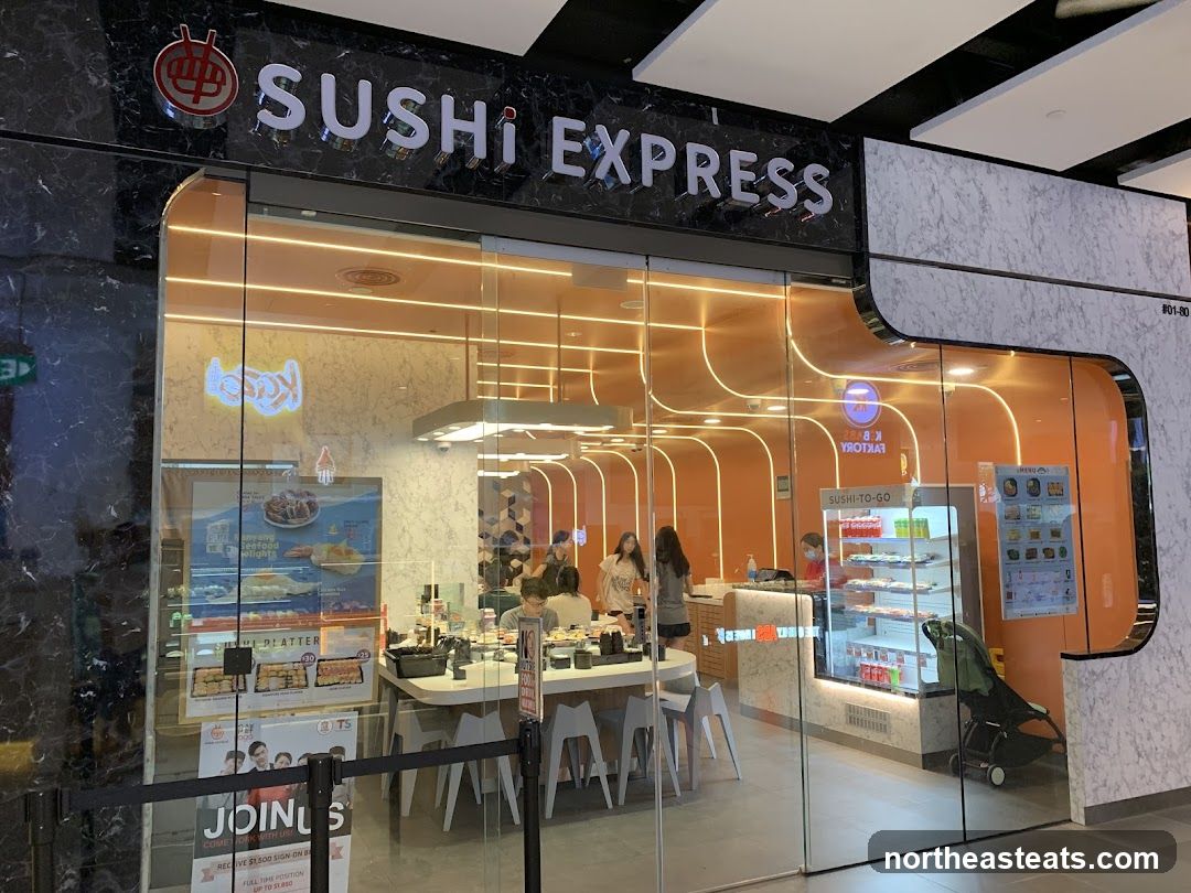 The exterior of a Sushi Express restaurant with large glass windows and doors. The restaurant's name is prominently displayed in large silver letters above the entrance. Inside, customers are seated at tables, and there is a display of sushi and drinks.
