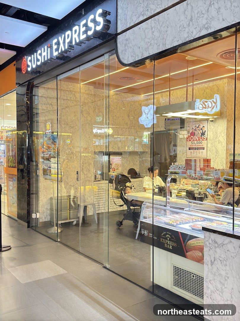 The exterior of a Sushi Express restaurant with large glass windows and doors. A prominent sign above the entrance reads "SUSHI EXPRESS". Inside, customers and staff are visible, along with displays of sushi and other food items.