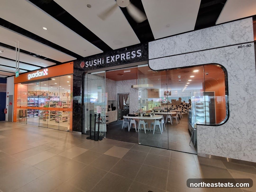 The exterior of two adjacent shops. The shop on the left is Guardian, a retail store with its orange signage and glass storefront displaying various products. The shop on the right is Sushi Express, a sushi restaurant with a glass entrance revealing the interior seating area and a display case. The signage for Sushi Express is visible above the entrance.