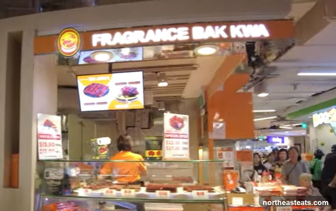 A brightly lit stall with a prominent sign reading "FRAGRANCE BAK KWA" above a display of meat products. Digital screens show images of the food, and price lists are visible. A person in an orange shirt is working behind the counter. Several customers are browsing in front of the stall.