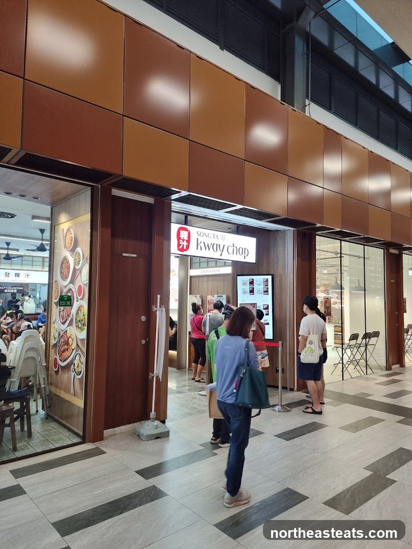 The exterior of a Song Fa Kway Chap restaurant. A sign with the restaurant's name is visible above the entrance. People are queuing outside the restaurant. A large menu board with pictures of food is displayed on the wall to the left of the entrance.