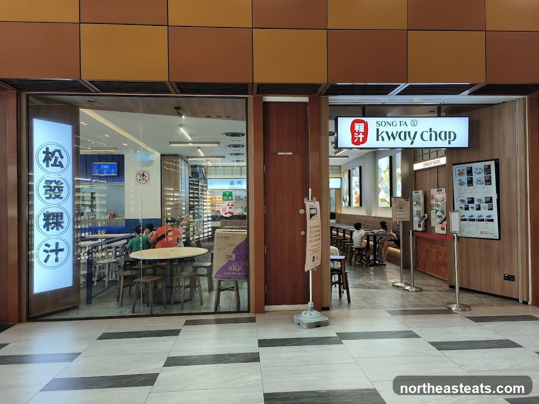 The exterior of a Song Fa Kway Chap restaurant. The left side of the storefront features a large glass window with Chinese characters and a circular logo. Inside, tables and chairs are visible, with patrons seated. The right side of the storefront has a prominent illuminated sign that reads "SONG FA kway chap" with a red logo. Below the sign, there is an "ORDER HERE" sign and a menu board displaying food items.