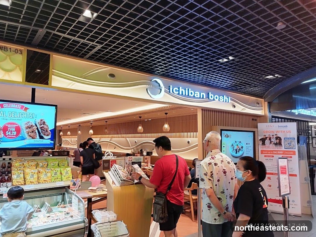 A view of the Ichiban Boshi restaurant entrance with customers. A digital screen displays a promotion for "Special Delights" with an offer of 2 for $4.20. The restaurant's name, "ichiban boshi," is visible on the facade. Another screen shows a recruitment advertisement. Shelves stocked with snacks are visible to the left.