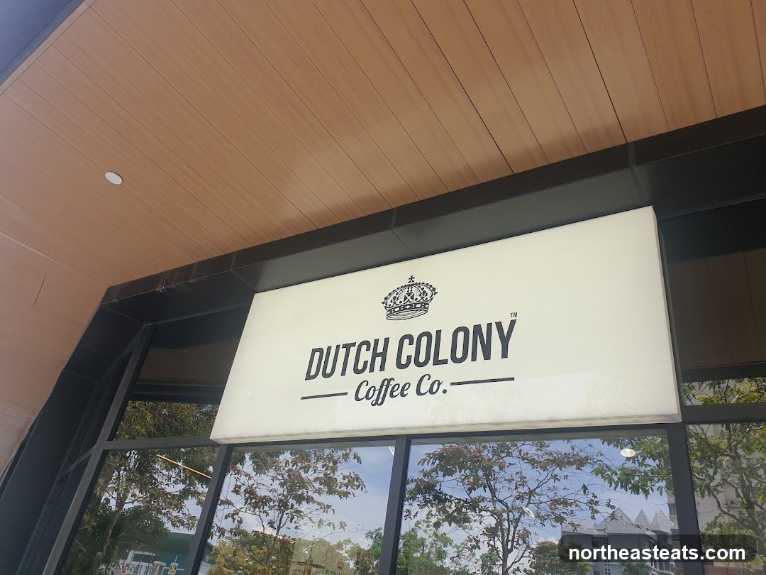 A lit sign with the words "DUTCH COLONY" and "Coffee Co." below it, along with a crown logo. The sign is mounted above a large window. The building has a wooden-paneled ceiling.