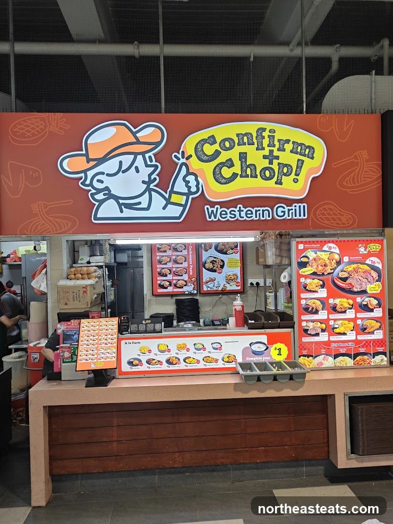A food stall with a large sign displaying the name "Confirm+Chop! Western Grill" and a cartoon character giving a thumbs up. Below the sign are menus with pictures of food items and prices. The stall appears to be in a food court.