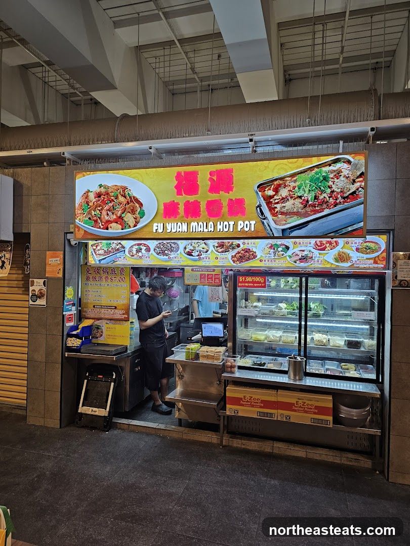 A food stall with a large illuminated sign displaying "FU YUAN MALA HOT POT" in English and Chinese. The sign features images of food. Below the main sign, there are smaller images of dishes and prices. A glass display case shows various ingredients. Two staff members are visible, one interacting with a customer and another preparing food. The stall is located in an indoor market or food court.