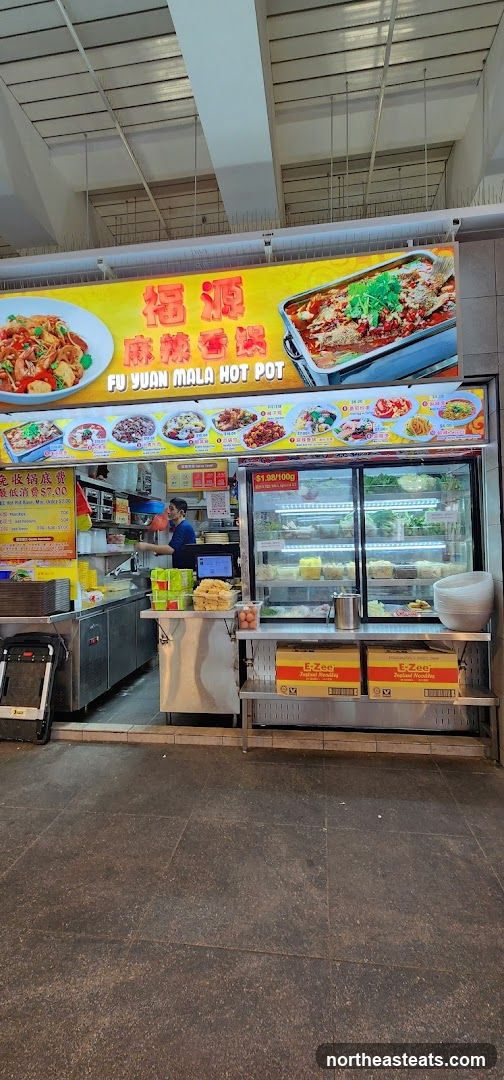 A brightly lit food stall with a large yellow sign displaying the name "FU YUAN MALA HOT POT" in English and Chinese, along with images of food. Below the main sign, a row of smaller images showcases various dishes. The stall has a counter with a display fridge filled with ingredients, and a staff member is visible working behind the counter. A price of "$1.98/100g" is displayed.