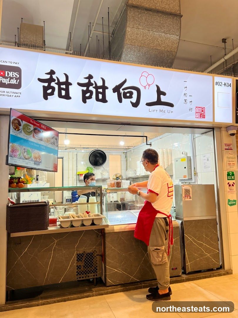 A dessert shop with a prominent sign displaying Chinese characters and the English name "LIFT ME UP". A menu board is visible to the left, showing "Hot Dessert" and "Cold Dessert" options. Two staff members are present, one serving a customer.