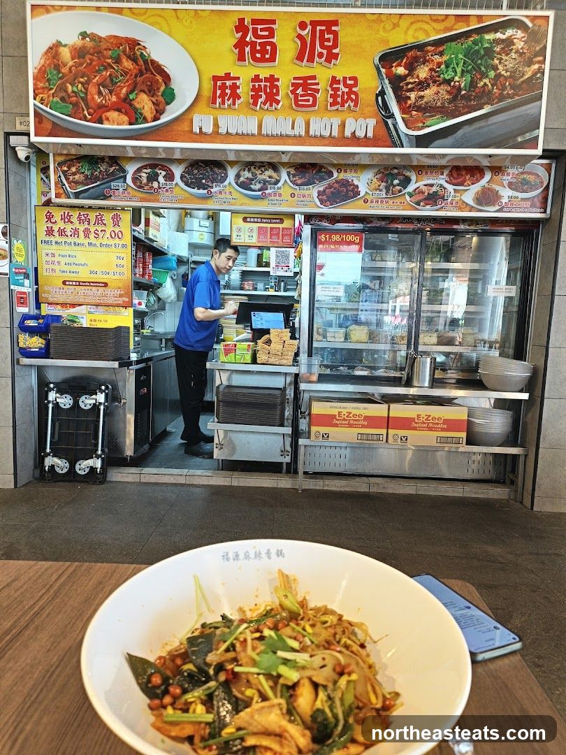A street-level view of a food stall with a prominent yellow sign displaying the business name and images of food. Below the main sign, a menu board shows various dishes with prices. A staff member in a blue shirt is visible behind the counter, interacting with equipment. The stall has glass display cases and shelves stocked with ingredients and products. In the foreground, a bowl of food is placed on a wooden table.