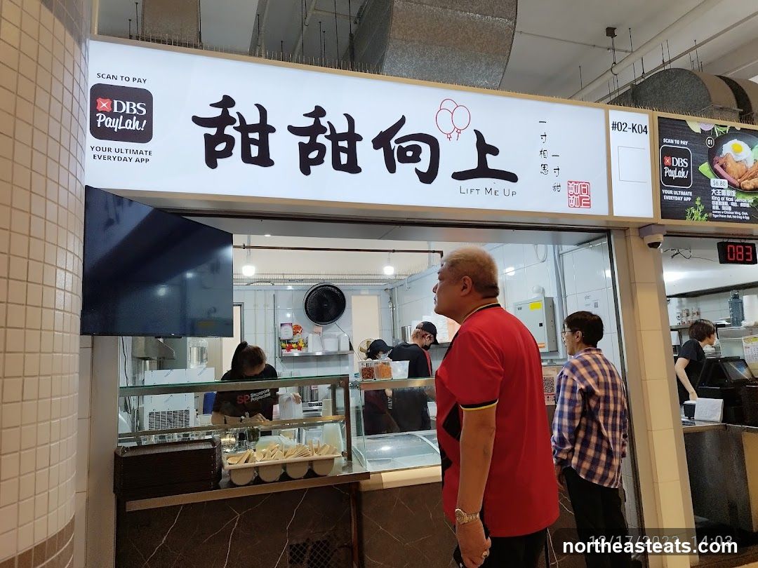 A food stall with a large illuminated sign displaying the name "甜甜向上 LIFT ME UP" in Chinese and English. The sign also features the DBS PayLah! logo and QR code for payment. Below the sign, the stall's interior is visible, with staff preparing food. Several customers are standing in front of the stall.