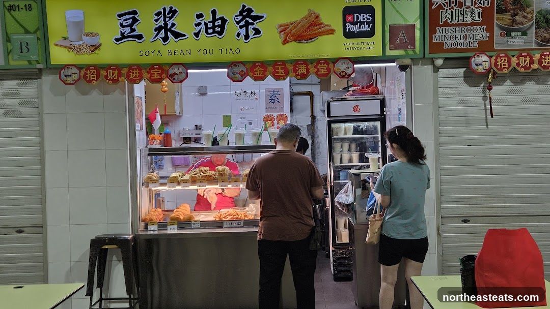 A food stall with a bright yellow sign displaying "SOYA BEAN YOU TIAO" in English and Chinese characters. Below the main sign, red octagonal decorations with Chinese characters hang. The stall displays various fried dough items and drinks in a refrigerated display case. Two customers are visible, one ordering and another looking at the offerings. The stall is part of a larger market or food court with shuttered stalls on either side.