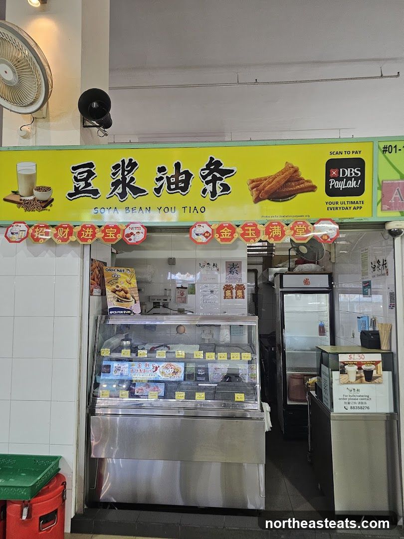 A storefront with a bright yellow sign featuring Chinese characters and the English text "SOYA BEAN YOU TIAO". Below the sign, a display case showcases various food items. To the right, a refrigerator is visible. Decorative banners with Chinese characters hang above the display.