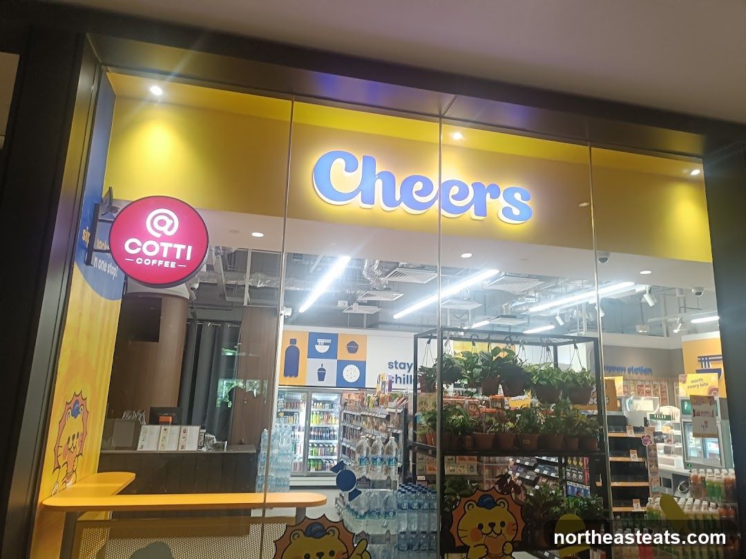 The exterior of a Cheers convenience store with a Cotti Coffee counter visible inside. The store has a bright yellow facade with the word "Cheers" illuminated in blue letters above the entrance. A circular red sign with "@ COTTI COFFEE" is displayed to the left of the entrance. Inside, shelves are stocked with drinks and snacks, and plants are displayed on hanging racks.