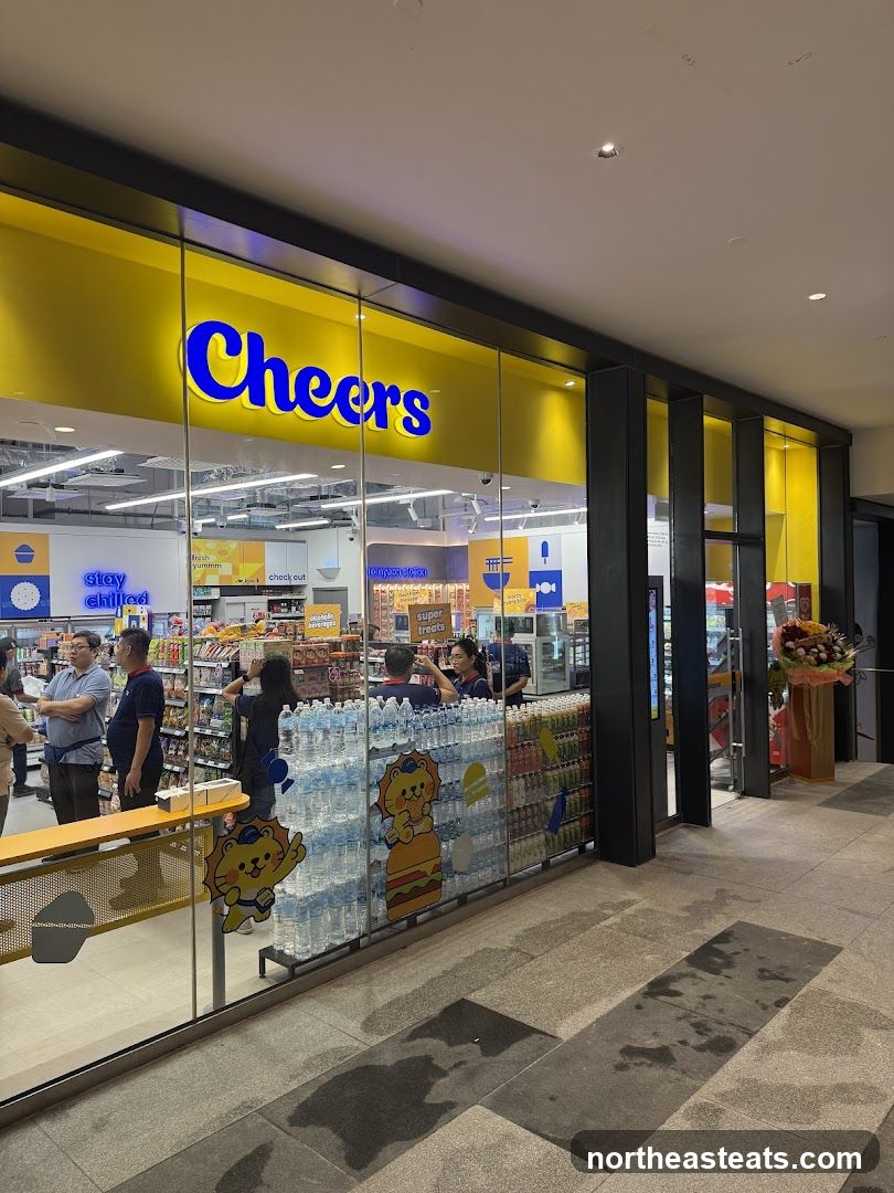 The exterior of a brightly lit convenience store with a yellow facade and large glass windows. The store's name, "Cheers," is prominently displayed in blue neon letters above the entrance. Inside, shelves are stocked with various products, and customers and staff can be seen. The windows are decorated with cartoon characters and displays of bottled water and other beverages.