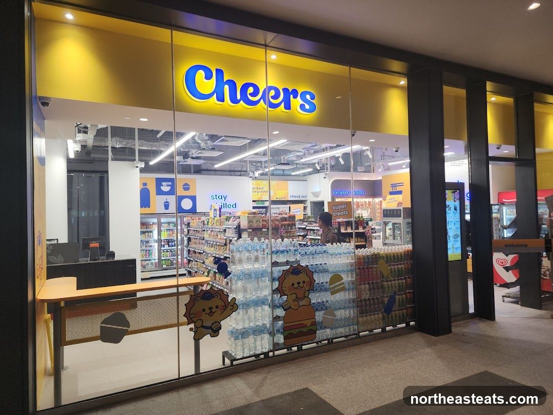 The exterior of a Cheers convenience store with a bright yellow awning and a large glass storefront. The store's name, "Cheers," is prominently displayed in blue lettering on the awning. Inside, shelves are stocked with various drinks and snacks. A counter with a computer is visible near the entrance. Decorative cartoon lions are placed on the glass.